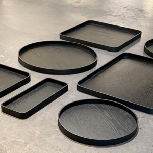 Collection of XLBoom Serve trays in sleek black finish, displayed in various shapes and sizes—round, rectangular, and square—on a smooth concrete floor. Each tray features a subtle wood grain texture and raised edges, showcasing the minimalist design and versatility of the Serve series.