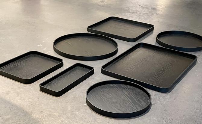 Collection of XLBoom Serve trays in sleek black finish, displayed in various shapes and sizes—round, rectangular, and square—on a smooth concrete floor. Each tray features a subtle wood grain texture and raised edges, showcasing the minimalist design and versatility of the Serve series.