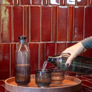 Two ribbed glass bottles and two ribbed tumbler glasses, one in clear glass and one in green, displayed on a round wooden Deck Tray D40. The set is placed against a glossy red tiled background, creating a warm and elegant composition. A hand pours the water in the glass.