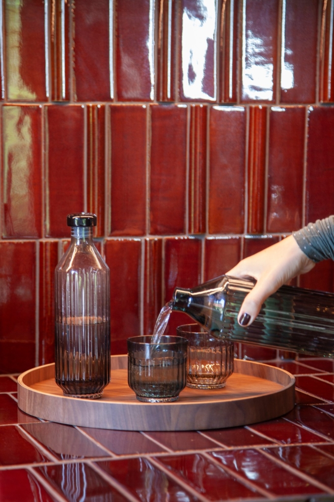 Two ribbed glass bottles and two ribbed tumbler glasses, one in clear glass and one in green, displayed on a round wooden Deck Tray D40. The set is placed against a glossy red tiled background, creating a warm and elegant composition. A hand pours the water in the glass.
