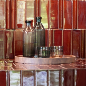 Two ribbed glass bottles and two ribbed tumbler glasses, one in clear glass and one in green, displayed on a round wooden Deck Tray D40. The set is placed against a glossy red tiled background, creating a warm and elegant composition.