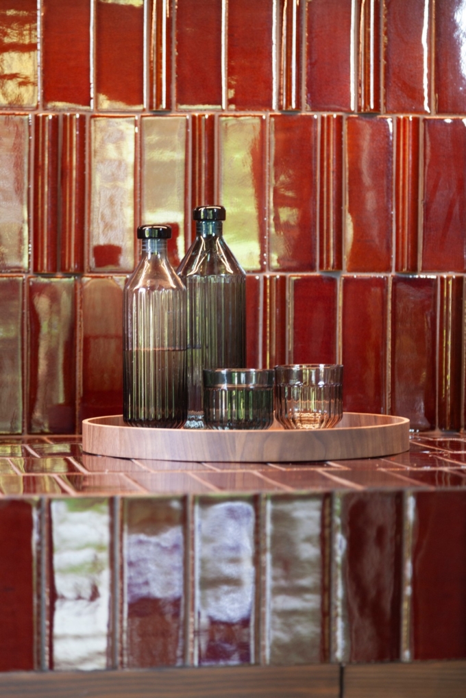 Two ribbed glass bottles and two ribbed tumbler glasses, one in clear glass and one in green, displayed on a round wooden Deck Tray D40. The set is placed against a glossy red tiled background, creating a warm and elegant composition.